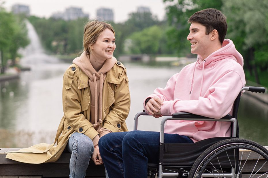 Young woman sitting on a bridge and talks with young man sitting in wheel chair.