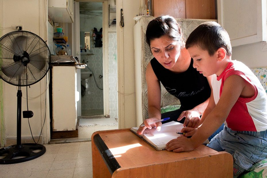 A mother sit besides her son as they complete work at a table in a run down apartment.