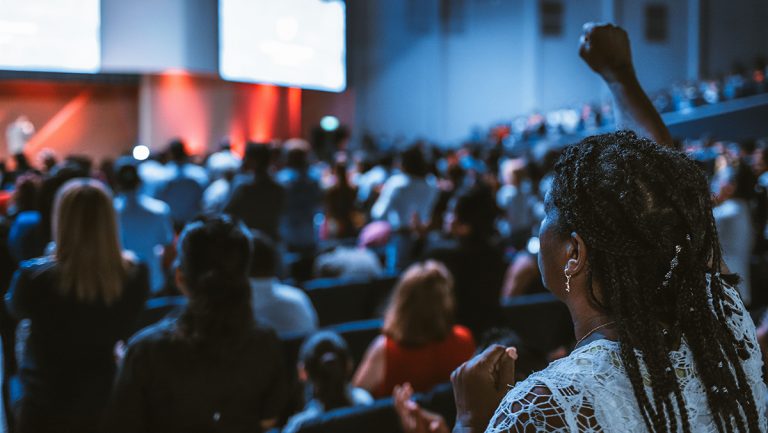 A woman stands up with her fist in the air in an auditorium full of people.