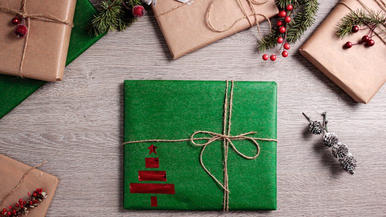 A view of a table from overhead. A present is wrapped with recycled green paper, brown rope thread and a red Christmas tree stamp.