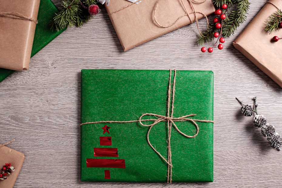 A view of a table from overhead. A present is wrapped with recycled green paper, brown rope thread and a red Christmas tree stamp.