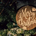 A close up picture of a section of a Christmas tree with a wooden ornament that says, "Merry Christmas".
