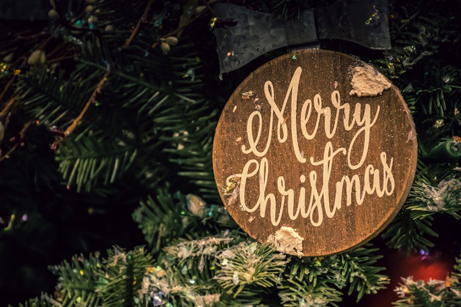 A close up picture of a section of a Christmas tree with a wooden ornament that says, "Merry Christmas".