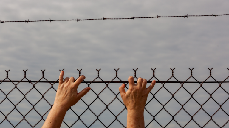 A pair of hands reaching up to climb over a barbed wire fence, with a with a dark grey sky